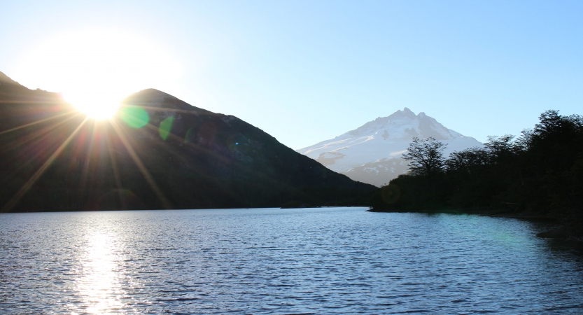 A blue body of water is framed by mountains and trees. In the background, there is a snow-covered mountain. 